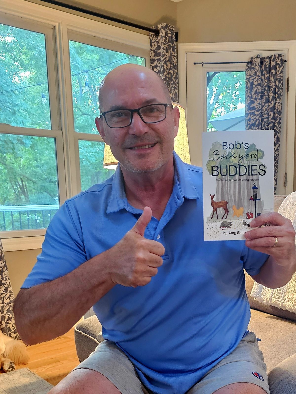Man in blue shirt holding a book titled 'Bob's Buddy Buddies' in a home setting.